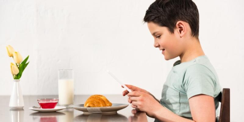 Un niño sentado ante una mesa con comida mira el teléfono móvil 