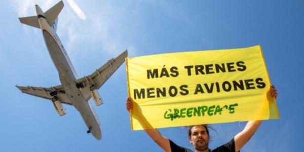 Un activista de Greenpeace sostiene un cartel en el que se lee 'Más trenes, menos aviones', con un avión sobrevolando de fondo 