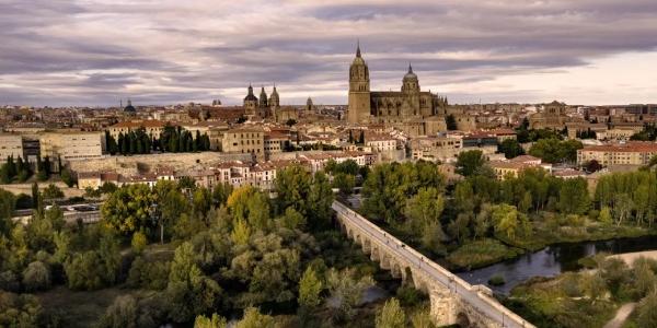 Vista panorámica de la ciudad de Salamanca al atardecer, mostrando la Catedral y el Puente Romano sobre el río Tormes, rodeados de vegetación.