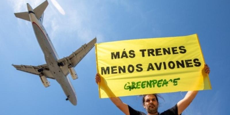 Un activista de Greenpeace sostiene un cartel en el que se lee 'Más trenes, menos aviones', con un avión sobrevolando de fondo 