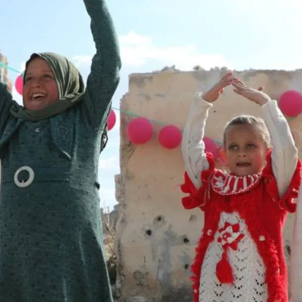 Tres niñas posan frente a un muro dañado en Ma'arrat An Nu'man, Siria. El muro está cubierto de agujeros de bala y adornado con globos rosas.