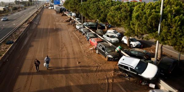 Dos personas caminan por la V-31 en Sedaví (Valencia) junto a decenas de coches apilados contra el quitamiedos, este jueves. 