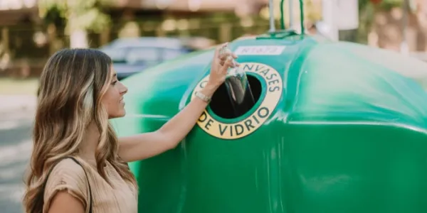 Una mujer depositando una botella de vidrio en el contenedor verde. 