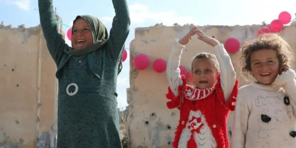 Tres niñas posan frente a un muro dañado en Ma'arrat An Nu'man, Siria. El muro está cubierto de agujeros de bala y adornado con globos rosas.